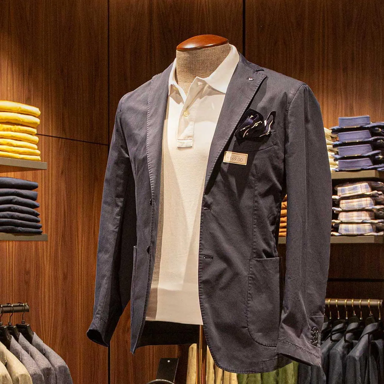 A mannequin dressed in a stylish navy blazer with a pocket square and a white polo shirt, displayed against a warm wooden background