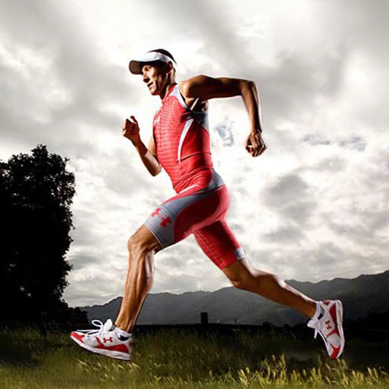 A man running outdoors in a vibrant red athletic suit and white visor, set against a backdrop of cloudy skies and distant mountains 